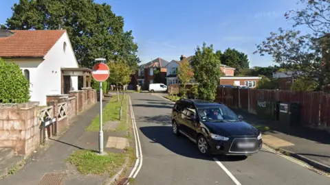 A car approaches a no-entry sign in Beaumont Road, which is narrow and tree-lined. A white church stands on the corner of the junction with the A36.