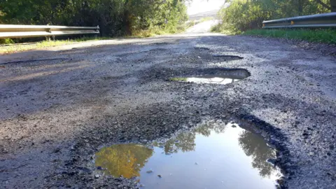 Getty Images Potholes in Tarmac on a road surface. They are filled with water. It is a sunny day
