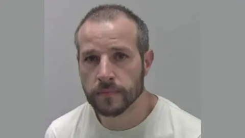 Police mugshot of a man with cropped dark hair and a short beard and moustache against a grey background.