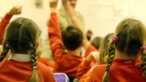 PA Media A shot from the back of a classroom. Children wearing orange jumpers are watching the teacher teach, whilst they raise their hands to answer questions.