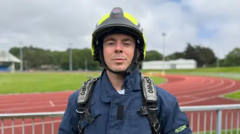 Nick Hays is wearing a blue fire fighter jacket, straps over his shoulders and yellow fluorescent helmet, standing in front of the orange tarmacked race track at the NSC.