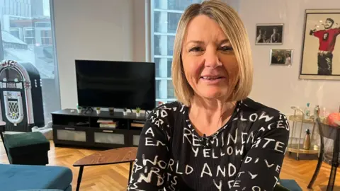 Nicky Wake is sitting on a chair in her living. In the background there are posters and photographs on the wall, a television and a jukebox. 
