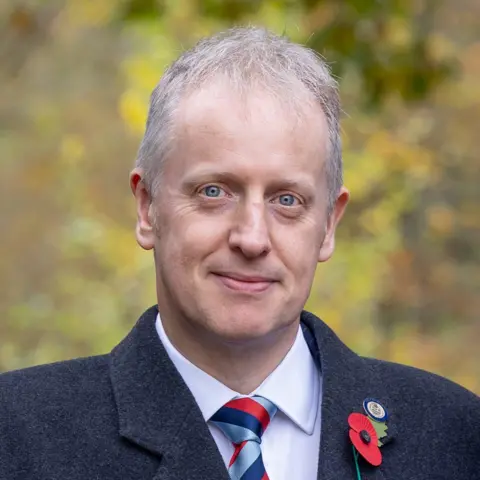 Rob Shenton A man with short white hear wearing a dark coat with a poppy pinned to it, with a white shirt and red and blue tie.