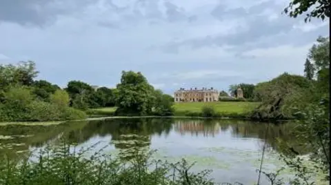 SOTTERLEY ESTATE A large, stately home nestled on a grassy knoll is framed by trees with a lake in the foreground 