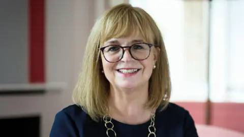 Lady Elish Angiolini looking directly at the camera. She is wearing dark-rimmed glasses and has shoulder-length light hair. She is wearing a dark top with a gold looped chain around her neck.