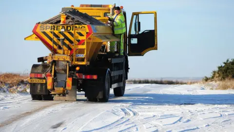 A gritter truck at work in North Yorkshire. The vehicle is stood on a snow-covered road with a man in yellow overalls stood on the side of the vehicle. 