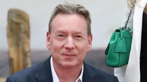 Frank Gardner. A man with blonde hair wearing a suit looking at the camera. There is a green hand bag to his left.