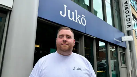 Shane McGann with light short hair and a white top with 'Juko'. He is standing in front of the cafe which also has the shop name on it over a blue background. The building is grey.