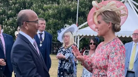 Hull University Teaching Hospitals NHS Trust A man in a dark suit speaking to a woman in a pink floral dress and hat at an outdoor formal event. Other guests in smart attire stand nearby on a grassy lawn, with a white marquee tent in the background.
