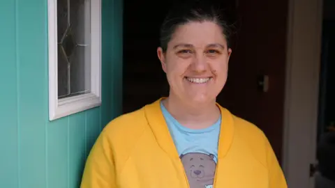Qays Najm/BBC Head and shoulders image of Kim Turner at her front door, which is turquoise. She has tied back dark hair and is wearing a yellow jumper with a blue t-shirt underneath with a bear on it.