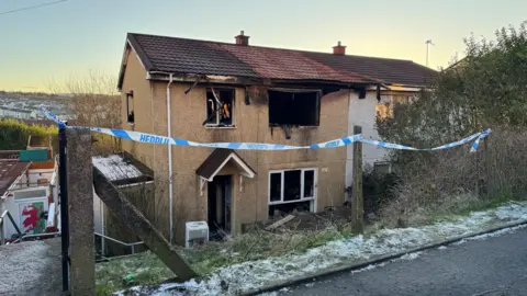 Burnt out end-of-terrace house with blackened windows and front door surrounded by police tape. The house is situated a little below street level with steps down. There is snow on the grassy verge above