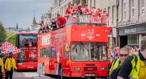 Adrian Thomson The Aberdeen men's football team atop a red open-top double decker bus with fans watching and cheering below in Aberdeen gear