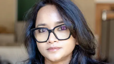 A woman - Prof Ronita Bardhan - looking directly at the camera with a neutral expression. She is wearing black glasses and has dark hair.