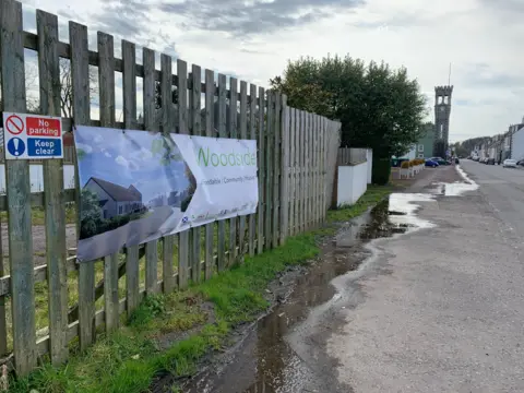 A fence with a developers sign on it and puddles in front, with a clock tower in the distance.