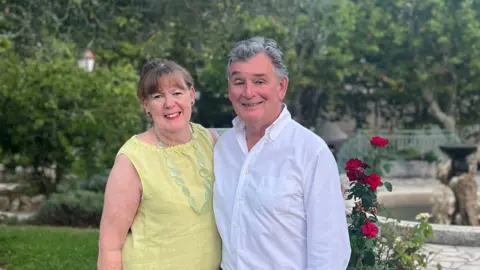 Family handout/PA A photo of Judy and Jonathan Bloomer. They are standing together with their arms around each other. They are standing in a garden with bushes and flowers behind them. Judy Bloomer is wearing a yellow top and Jonathan Bloomer is wearing a long-sleeved white shirt.