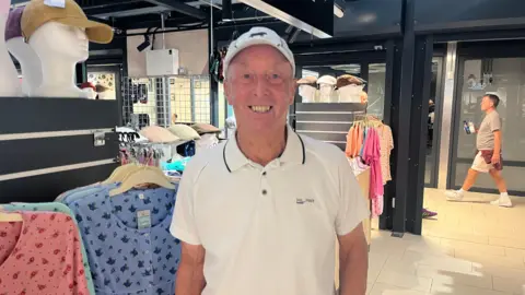 Man wearing white polo shirt and cap smiling in front of a clothes stall.