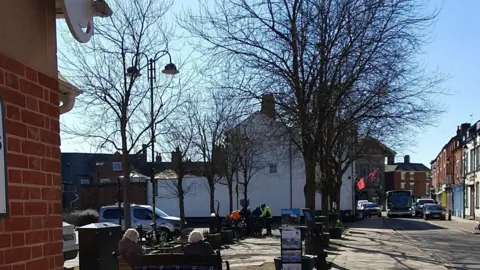 Two lines of large trees on a promenade-style path in a town. There are people sitting at a bench looking towards them. A road is to the right hand side with vehicles driving towards the camera