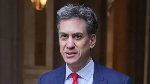 PA Media Ed Miliband is pictured smiling at the camera. He has short black and grey hair. He is wearing a dark blue suit with a light blue shirt and red tie.