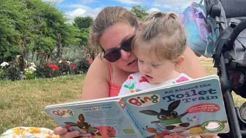 The mum is reading a book called 'all aboard the toilet train' to her daughter. The daughter is looking at the book and sat on the mum's lap and she is wearing sunglasses 