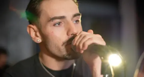 Jamie Niblock/BBC Lead singer Ben has black facial hair, and is gripping on to the microphone as he sings into it.