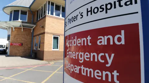 A sign saying St Peter's Hospital in black writing, with a red block underneath with white text saying accident and emergency department. Behind is a brock building. 