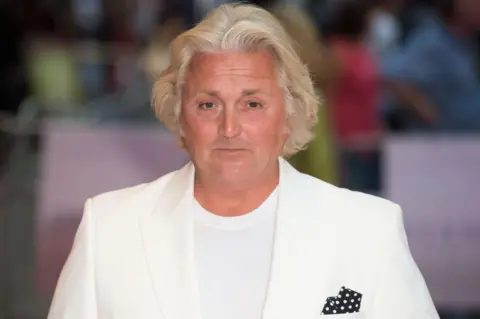 Getty Images Head and shoulders shot of David Emanuel. He has white hair and looks at the camera. He is wearing a white t-shirt and white shirt with a black and white polka-dot scarf in the left-hand pocket.