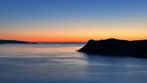 A sunset with a body of water in the foreground and a hill to the right of the photo