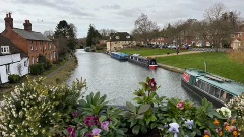 Solentgirl A canal runs through the centre of the picture with three barges on the right-hand side. In the foreground there is a flower box full of brightly coloured flowers and beyond the barges are stretches of green grass lined by cars and houses. There are also a few houses on the left-hand side of the canal.