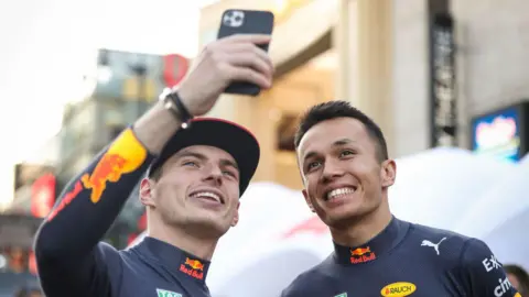 Getty Images Max Verstappen takes a selfie with Alex Albon while they were Red Bull teammates. Max is on the left and is holding a phone up in the air with his right hand to take the selfie. Both men are smiling and both are wearing navy blue Red Bull long sleeve racing tops.