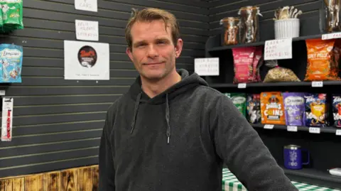 Shaun Campbell standing behind the counter of his pet supplies stall. He has short blond hair and is wearing a black jumper.