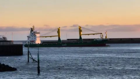 Martin Emmerson/BBC A large cargo ship which is white, blue and yellow. The front of the ship is slightly higher in the water than the back. The sea looks calm and there is a long pier behind it and what looks like another ship to the left. The sun is still rising and the sky is blue and yellow.