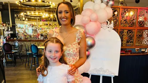 A young girl in a white dress, and her mother in a flowery dress, stand in front of pink, silver and white balloons. In the background there is a large chandelier hanging over a bar, with tables and chairs. 