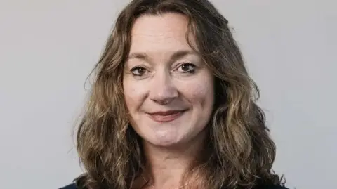 TYNWALD Daphne Caine, who has long, wavy, light brown hair, against a grey background.