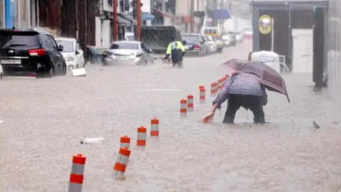 A man, hunched over and holding a polka-dotted umbrella, makes his way through a flooded street, caused by torrential rain, in Gwangju, South Korea on 17 July, 2025. A row of vehicles are parked on the left of the street. The floodwaters have submerged parts of the vehicles' wheels. 