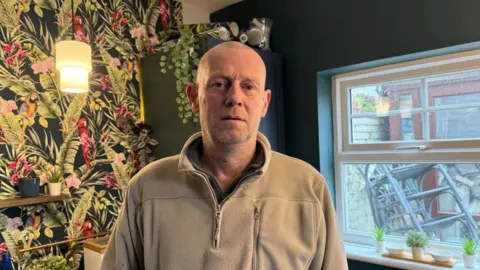 Andrew Humphries stands in a cream fleece in front of a garden wallpaper with parrots on it and leaves - a window over his left shoulder has stacked chaits in it. There are plants on the windowsill. 