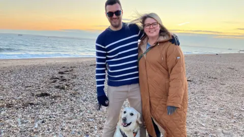 Laura Johnston stands next to her husband Colin on Southsea seafront. The pebbled beach and the sea can be seen in the background, with the start of an orange sunset in the distance. Colin stands on the left, he is wearing a blue knit jumper with white stripes and cream chinos, with black thick framed sunglasses and black gloves. He has his arm around Laura, standing to the right. She has blond hair and wears red thick framed glasses and a mustard yellow puffer duvet coat and a blue and white striped top underneath. Their dog Willow, a fully grown white golden retriever, is sat in between Colin's legs with her tongue sticking out like she's panting. They are all look at the camera and smiling.