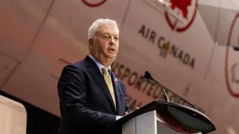 Getty Images An image showing Michael Rousseau at a podium, standing in front of an Air Canada plane that has the airline's red maple leaf logo next to the Olympics logo. He has gray hair, and is wearing a dark navy suit and a yellow patterned tie.