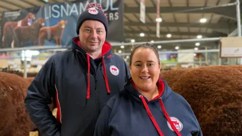 BBC A man and woman in branded navy and red hoodies stand inside a livestock hall in front of cattle, with a banner reading “Lisnamul Farmers” visible in the background. 