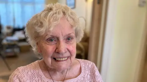 A picture of Shirley smiling at the camera. She is wearing a necklace and a pink shirt.