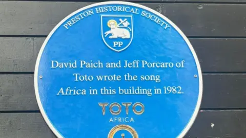 All Hopes No Promises Blue plaque picture, which reads that song Africa by Toto was written in Preston - in the building that is now a burger restaurant, in 1982. The plaque is handed on a black wall.