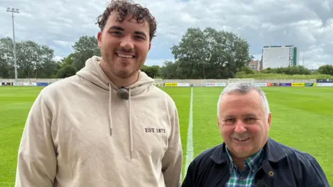 A man in a beige hoodie stands with a man in a blue jacket and checked shirt, both smile into the camera