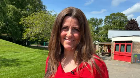 Cathy Killick/BBC A woman with long brown hair wearing a red top stands in a school playground. She is smiling at the camera