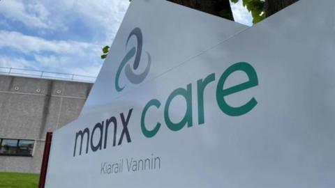A close-up of a sign that says Manx Care. The sign is white with grey and green lower case letter and the Manx translation is written in smaller grey lettering below it.