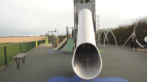 A new play area including a metal slide, rubber flooring and a range of apparatus. People can be seen playing and grey skies behind.