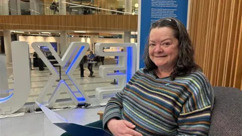 A woman in a multi coloured striped top smiles. She has dark grey / brown hair and sits in a grey chair. Behind her is a wooden panel.