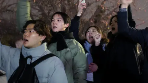 Reuters Young people in puffer jackets hold their fists and phones in the air as they shout slogans while protesting outside the National Assembly on Tuesday