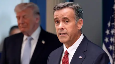 Ratcliffe looks at the camera while speaking with Trump out-of-focus in the background next to a US flag, pictured in the White House in April.