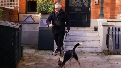 PA Media Gary Lineker in a dark coat walking his dog outside his red brick home in London