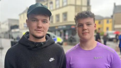 Cory McLean, right, looking right at the camera. He is wearing black, fleeced Nike hoodie with the Nike logo in the top left corner. He is wearing a dark green cap. Taylor Montgomery, left, looking right at the camera. He is wearing a light purple t shirt with a Nike logo in the top left corner.