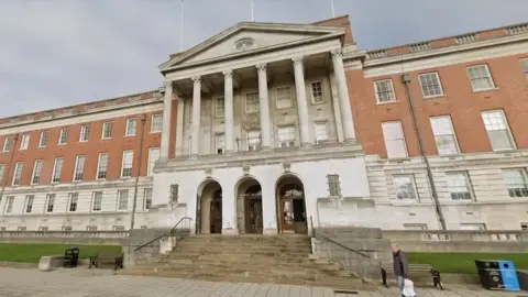 Google An imposing looking council office building in red and white stone with six columns above a triple doorway and a wide stairway from the pavement below